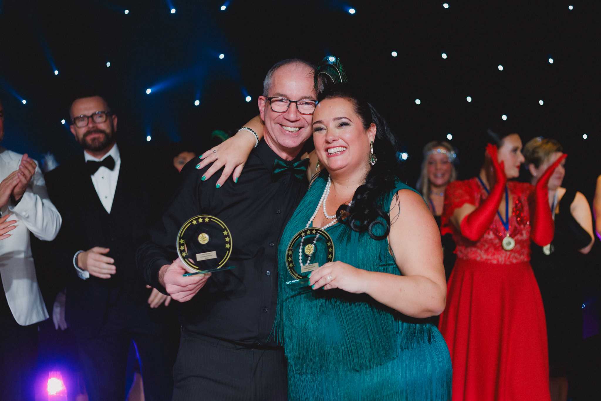 Winners Diane in a green dress and Dave in a black suit standing holding the winning trophies